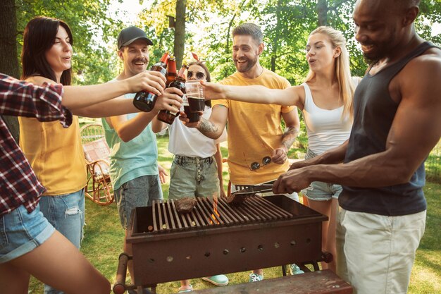 Groupe d'amis heureux ayant de la bière et un barbecue en journée ensoleillée. Repos ensemble en plein air dans une clairière ou une cour