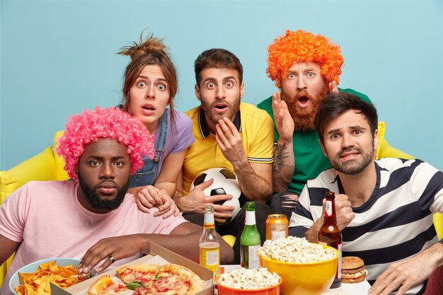 Un groupe d'amis multiethniques regarde, a retenu son souffle tout en regardant un match de football très excitant, asseyez-vous sur un canapé près de la table avec une pizza, de la bière et du pop-corn isolé sur un mur bleu. Réaction émotionnelle folle