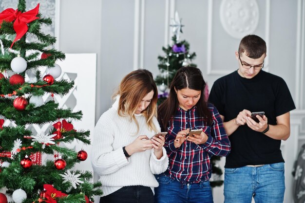 Groupe d'amis tels que deux filles et un homme regardant des téléphones portables contre l'arbre de noël en studio