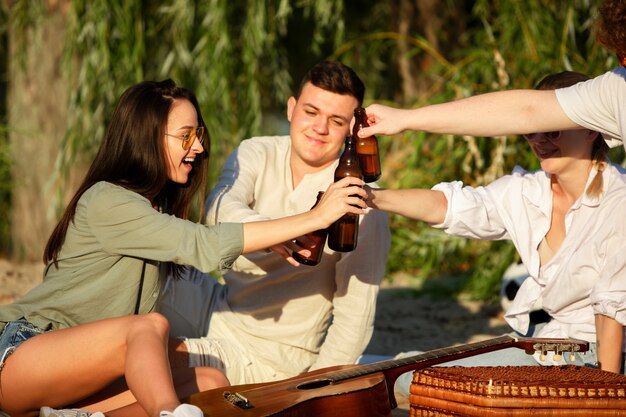 Groupe d'amis tinter des bouteilles de bière pendant un pique-nique à la plage. Mode de vie, amitié, amusement, week-end et concept de repos.