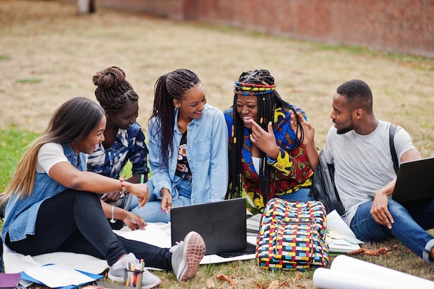 Groupe de cinq étudiants africains passant du temps ensemble sur le campus de la cour de l'université amis afro noirs assis sur l'herbe et étudiant avec des ordinateurs portables