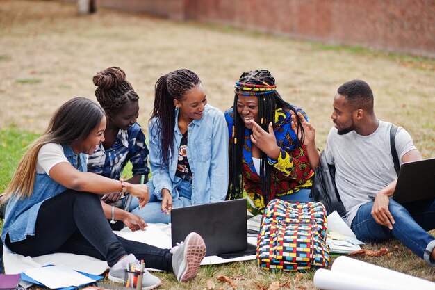 Groupe de cinq étudiants africains passant du temps ensemble sur le campus de la cour de l'université Amis afro noirs assis sur l'herbe et étudiant avec des ordinateurs portables