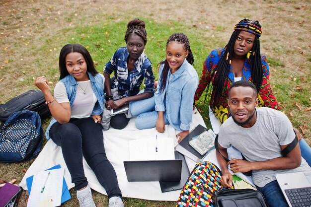 Groupe de cinq étudiants africains passant du temps ensemble sur le campus de la cour de l'université Amis afro noirs assis sur l'herbe et étudiant avec des ordinateurs portables