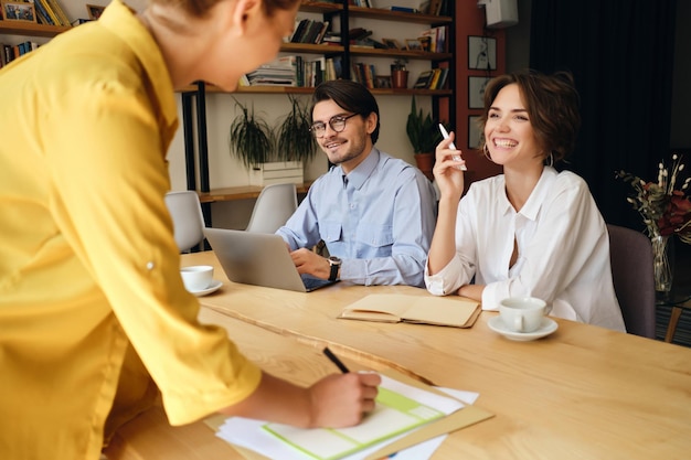 Groupe de collègues de travail joyeux assis au bureau avec un ordinateur portable et des papiers tout en parlant joyeusement au travail dans un bureau moderne