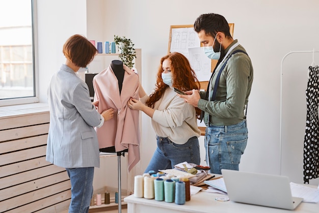 Groupe de créateurs de mode avec des masques médicaux travaillant en atelier et vérifiant le vêtement sur la forme de la robe