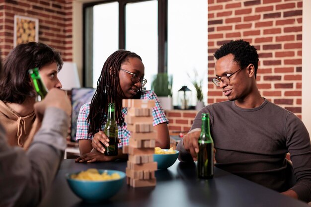 Groupe diversifié d'amis proches assis à table dans le salon tout en s'amusant avec des jeux de société. Des gens occasionnels assis à la maison tout en profitant d'activités de loisirs amusantes avec des blocs de bois