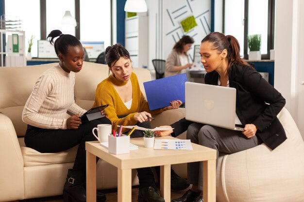 Groupe diversifié de femmes d'affaires assises sur un canapé dans le bureau d'une entreprise de démarrage d'entreprise moderne parlant de la gestion de projets financiers et de stratégies de démarrage, à l'aide d'un ordinateur portable et de graphiques