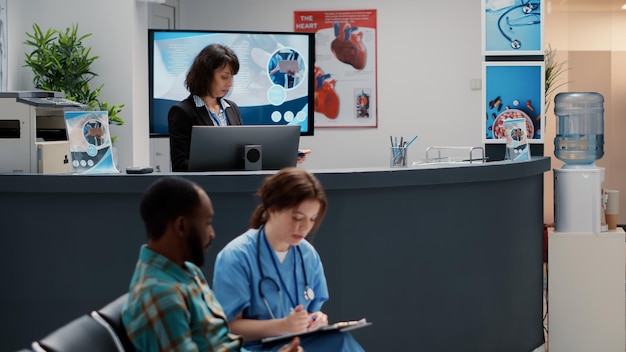 Groupe diversifié de patients demandant de l'aide à la réception de l'hôpital, assis dans le hall de la zone d'attente. Mère avec enfant, homme âgé et patient asiatique ayant rendez-vous dans une clinique médicale très fréquentée.