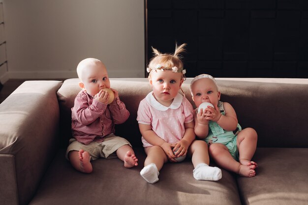 Groupe d'enfants mignons drôles assis ensemble sur un canapé jouant avec des boules colorées. Trois enfants heureux garçon et fille dans des vêtements colorés posant sur un canapé confortable plein coup