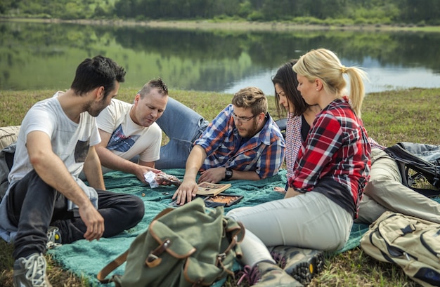 Photo gratuite groupe de jeunes amis assis sur l'herbe près du lac par une journée ensoleillée