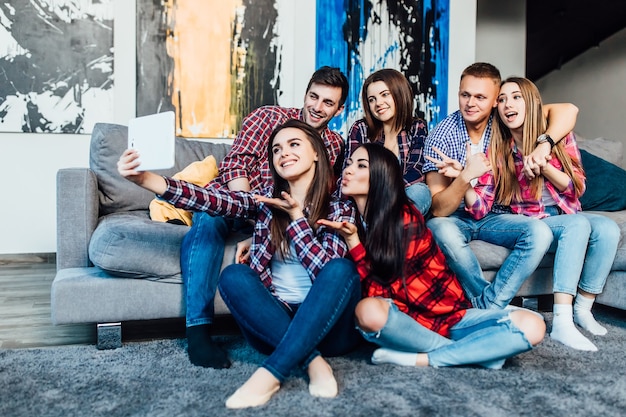 Groupe de jeunes amis drôles assis à la maison sur un canapé et faisant des selfies ensemble..