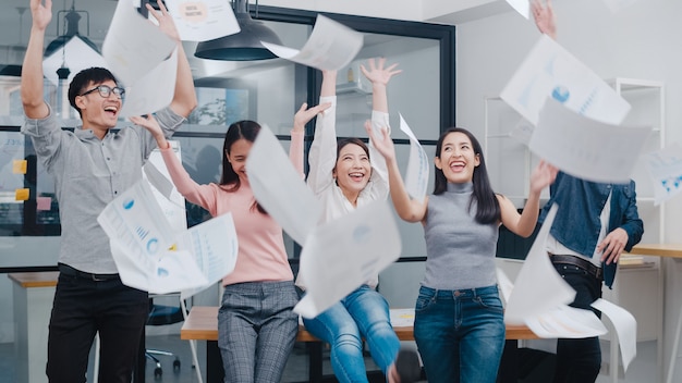 Un groupe de jeunes créatifs asiatiques en tenue décontractée et élégante célèbre le succès du projet et lance des documents au bureau.