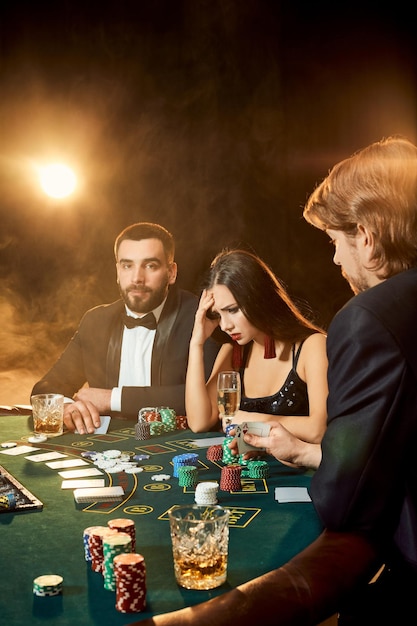 Un groupe de jeunes riches joue au poker dans le casino. Deux hommes en costume et une jeune femme en robe noire. Fumée. Casino. Poker