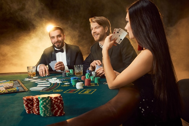 Un groupe de jeunes riches joue au poker dans le casino. Deux hommes en costume et une jeune femme en robe noire. Fumée. Casino. Poker