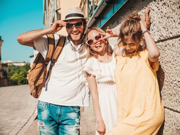 Groupe de jeunes trois amis élégants posant dans la rue. Homme de mode et deux jolies filles vêtues de vêtements d'été décontractés. Sourire, modèles, amusant, lunettes soleil., Gai, femmes, et, type, dehors