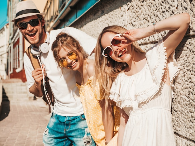 Groupe de jeunes trois amis élégants posant dans la rue. Homme de mode et deux jolies filles vêtues de vêtements d'été décontractés. Sourire, modèles, amusant, lunettes soleil., Gai, femmes, et, type, dehors