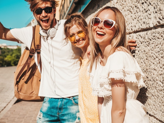 Groupe de jeunes trois amis élégants posant dans la rue. Homme de mode et deux jolies filles vêtues de vêtements d'été décontractés. Sourire, modèles, amusant, lunettes soleil., Gai, femmes, et, type, dehors