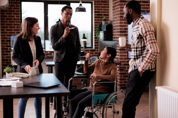 Groupe multiethnique de collègues riant dans un bureau d'affaires avec une femme paralysée assise en fauteuil roulant. Employé asiatique souffrant d'un handicap chronique et d'une déficience s'amusant avec ses collègues.