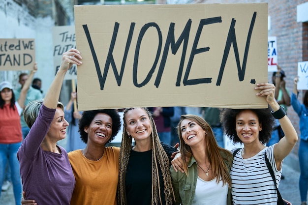 Groupe multiethnique de femmes heureuses tenant une bannière avec l'inscription "femmes" tout en participant à une manifestation pour les droits des femmes