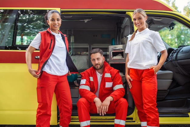 Un groupe multiethnique de trois ambulanciers paramédicaux à l'arrière d'une ambulance grimpant par les portes ouvertes Les deux femmes sourient à la caméra et leur collègue masculin a une expression sérieuse