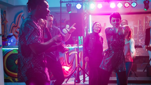 Groupe de personnes profitant d'une soirée disco, dansant sur la musique du club lors d'une réunion sociale. Jeunes hommes et femmes montrant des mouvements de danse sur la piste de danse, faisant la fête avec les lumières de la scène. Tir à la main.