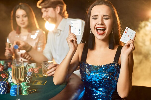 Groupe d'un peuple élégant jouant au poker à la maison de jeu. Concentrez-vous sur une brune émotionnelle dans une robe bleue brillante. Passion, cartes, jetons, alcool, dés, jeux d'argent, casino - c'est du divertissement. Danger