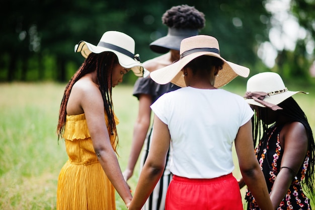 Groupe de quatre magnifiques femmes afro-américaines portant un chapeau d'été se tenant la main et priant à l'herbe verte dans le parc