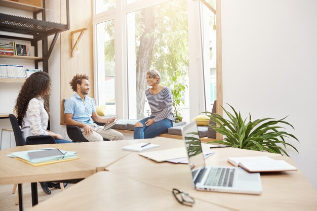 Groupe de trois jeunes entrepreneurs travaillant ensemble sur la recherche dans une bibliothèque lumineuse, discutant des idées de projets, parcourant la documentation, passant une matinée productive. Concept d'entreprise