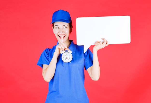 Guide féminin en uniforme bleu tenant un panneau d'information rectangulaire blanc avec un réveil et demandant le silence.