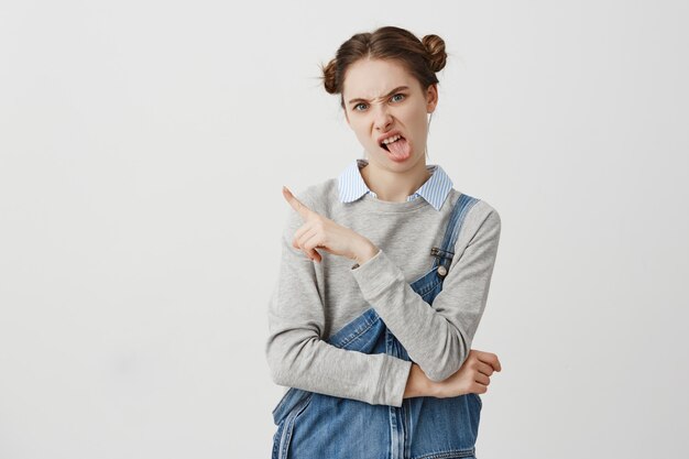 Guillerette femelle adulte montrant le doigt sur le mur blanc. Femme confiante avec double chignon mettant la langue dehors et exprimant ne pas aimer son choix. Espace copie