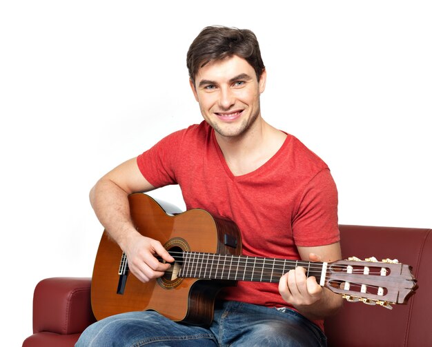 Le guitariste souriant joue sur isoler guitat acoustique sur blanc. Beau jeune homme est assis avec une guitare sur un divan