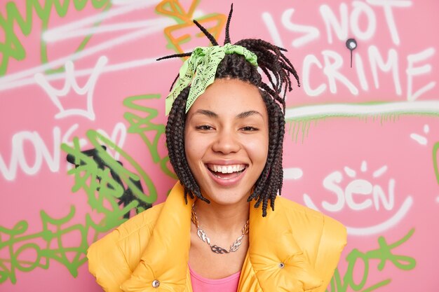 Gurl hipster positif sourit largement aime les poses de temps libre contre le mur de graffitis colorés vêtus de vêtements de style urbain décontracté étant de bonne humeur