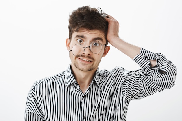 Guy après la gueule de bois a perdu la mémoire. Homme malpropre interrogé confus en chemise rayée, se gratter les cheveux et à l'ignorance et sans aucune idée avec un coup d'œil sous les lunettes, debout sur un mur gris