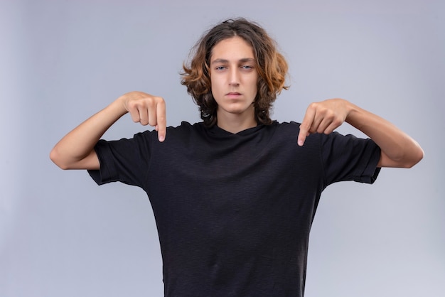 Photo gratuite guy aux cheveux longs en t-shirt noir pointe les doigts vers le bas sur le mur blanc