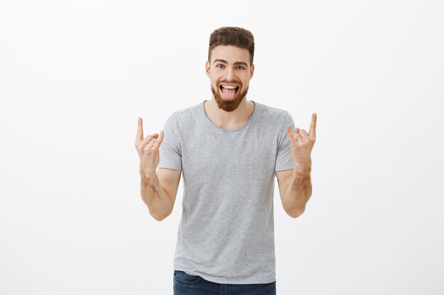 Guy à bascule à la fête apporte des vibrations positives. Insouciant surpris et heureux beau mâle charismatique caucasien avec barbe, yeux bleus montrant la langue montrant le geste de rock-n-roll bénéficiant de la musique cool en club