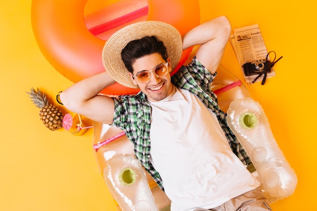 Photo gratuite guy en chapeau de paille, lunettes et t-shirt blanc repose sur un matelas gonflable avec cocktail d'ananas et appareil photo rétro.