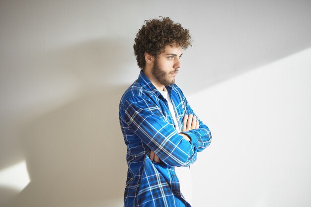 Guy hipster branché sérieux avec barbe floue croisant les bras sur sa poitrine en signe de désapprobation ou de réticence, debout isolé au mur blanc. Expression faciale humaine et langage corporel