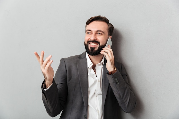 Guy positif en affaires en tenue de chat mobile portant des gestes lors de l'utilisation de téléphone portable, isolé sur gris