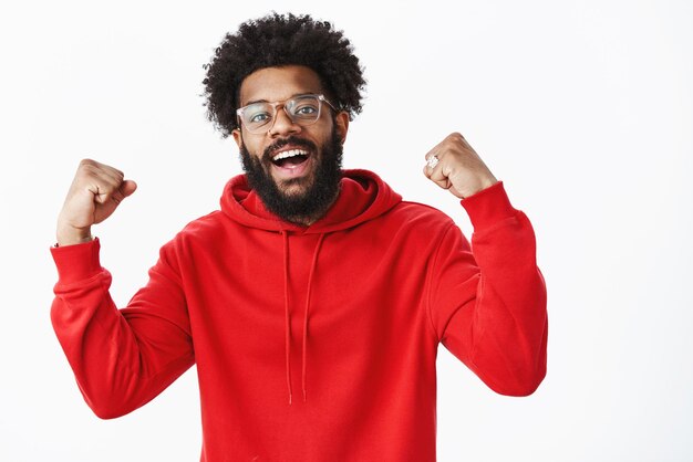 Guy se sentant génial gagner célébrant la victoire levant les poings dans le triomphe et le succès souriant largement comme de bonne humeur debout optimiste en sweat à capuche rouge sur fond gris obtenant un excellent résultat