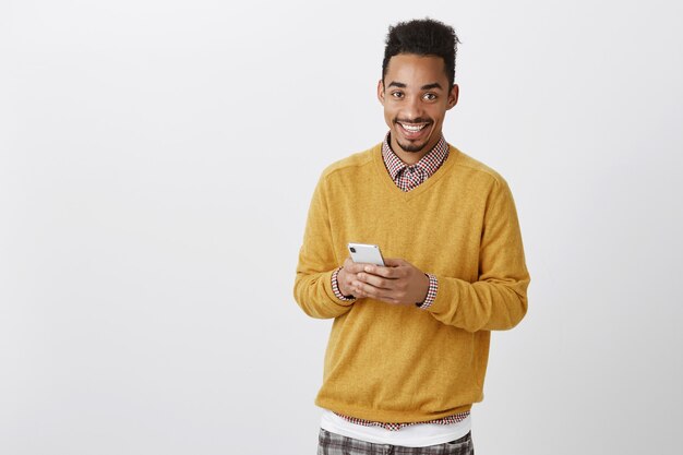 Guy veut passer un appel. Tir à l'intérieur d'un beau modèle masculin afro-américain heureux avec coupe de cheveux afro en pull jaune, tenant un smartphone, souriant largement tout en envoyant des messages avec un ami