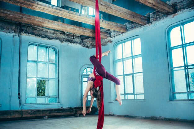 Gymnaste gracieuse effectuant des exercices aériens avec des tissus rouges sur fond bleu vieux loft. Jeune fille caucasienne de l'adolescence. Le cirque, acrobatique, acrobate, interprète, sport, fitness, concept de gymnastique