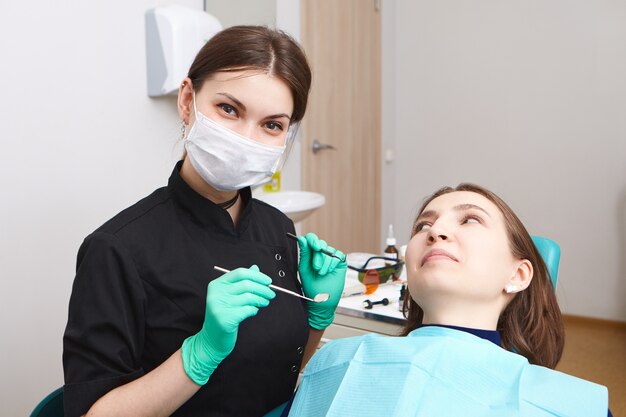 Habile confiant jeune dentiste femme portant des gants d'examen et un masque blanc tenant une sonde métallique et un miroir dentaire, prêt à examiner la cavité buccale d'une patiente assise dans une chaise