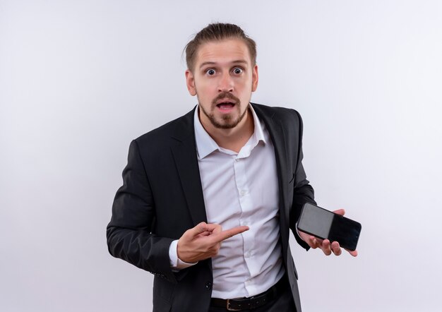 Handsome business man wearing suit holding smartphone pointant avec le doigt sur elle à la surprise et étonné debout sur fond blanc