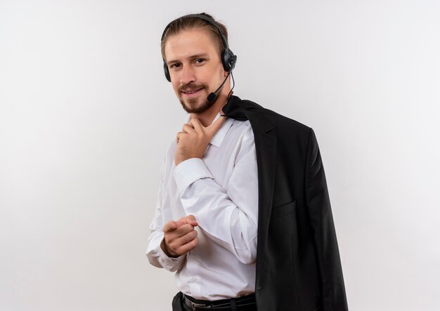 Handsome businessman holding jacket sur l'épaule avec un casque avec un microphone regardant la caméra avec un sourire confiant debout sur fond blanc