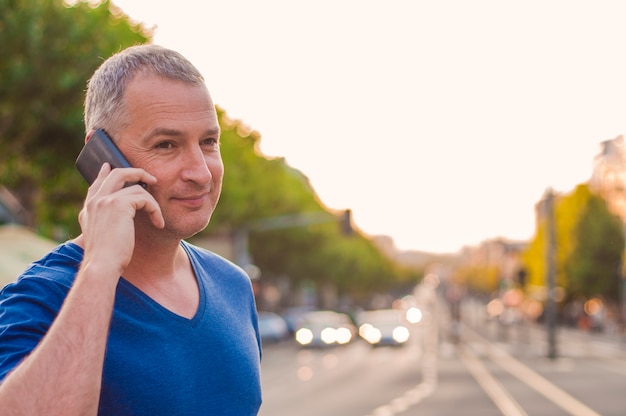 Handsome man tenant le téléphone mobile, utilisant un téléphone intelligent, téléphoner, parler au téléphone, se tenir sur une rue ensoleillée avec le trafic de transport en arrière plan