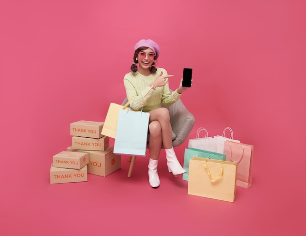 Happy Asian teen woman sitting on sofa holding shopping bags et smartphone isolé sur fond rose Shopper ou concept accro du shopping