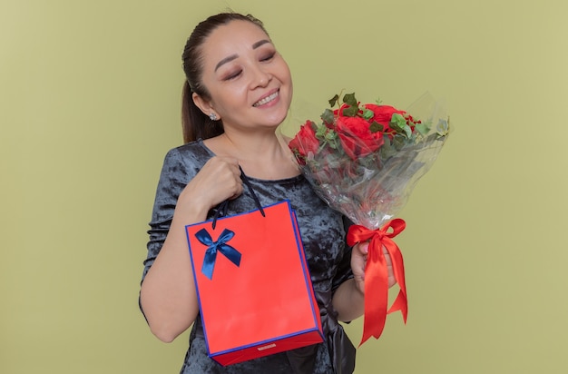 Happy Asian woman holding bouquet de roses rouges souriant joyeusement et sac en papier avec cadeau célébrant la journée internationale de la femme debout sur le mur vert