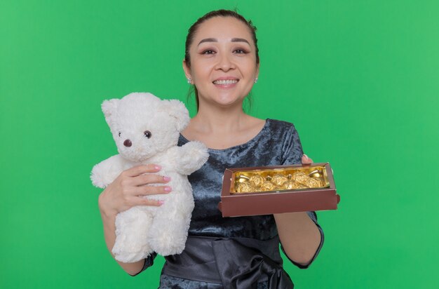 Happy Asian woman holding teddy bear et fort de bonbons au chocolat comme cadeaux souriant joyeusement célébrant la journée internationale de la femme debout sur le mur vert