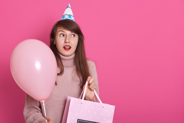 Happy brunette girl with pink balloon and bithday present in hands, garde la bouche ouverte, étant surpris, regarde de côté avec astin