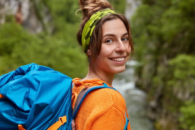 Happy girl traveller a un voyage d'exploration, porte un sac à dos, se tient à côté du ruisseau, sourit largement